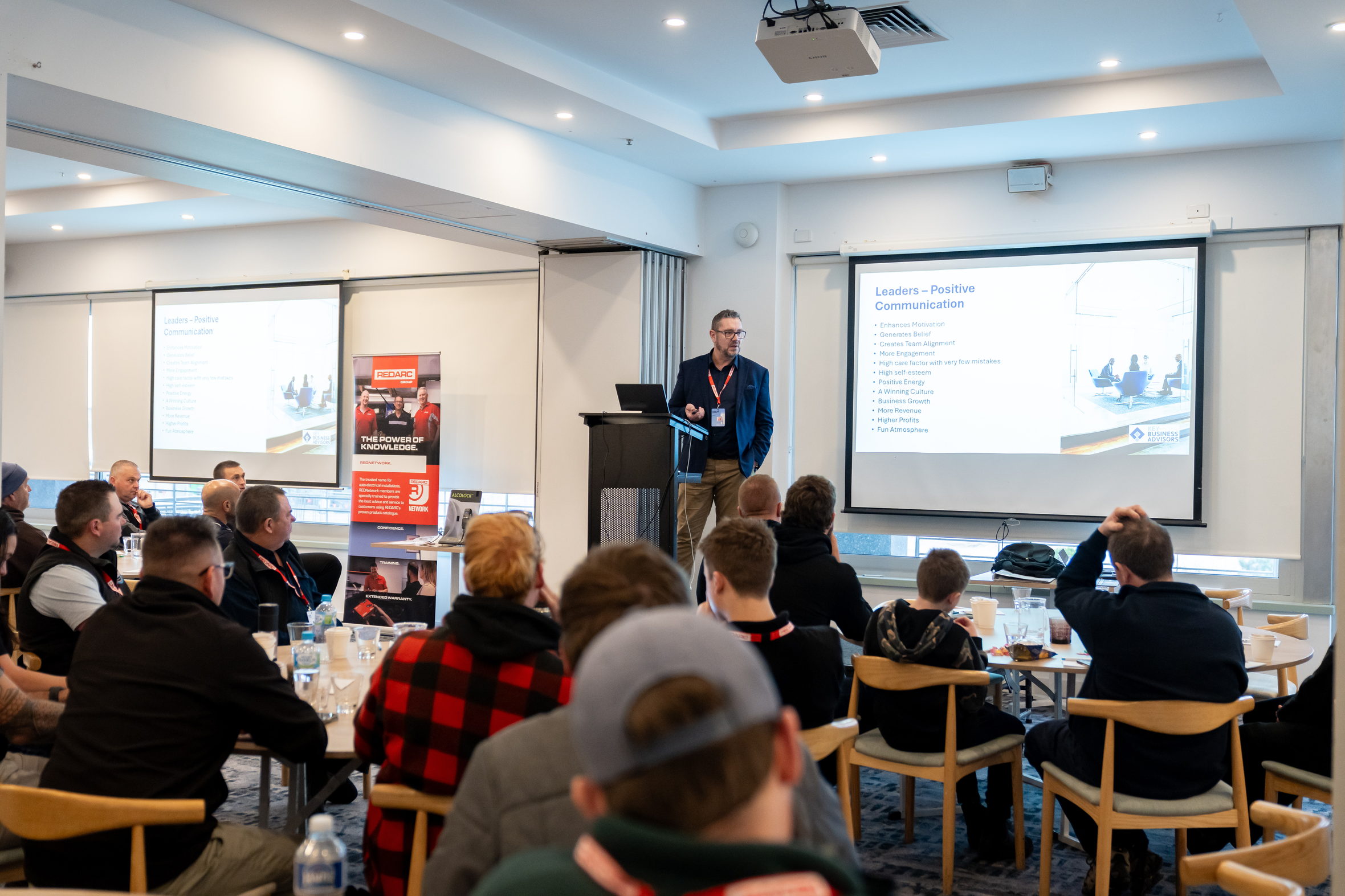 Colin Wilson delivering a leadership communication session to a seated audience, slides either side of him