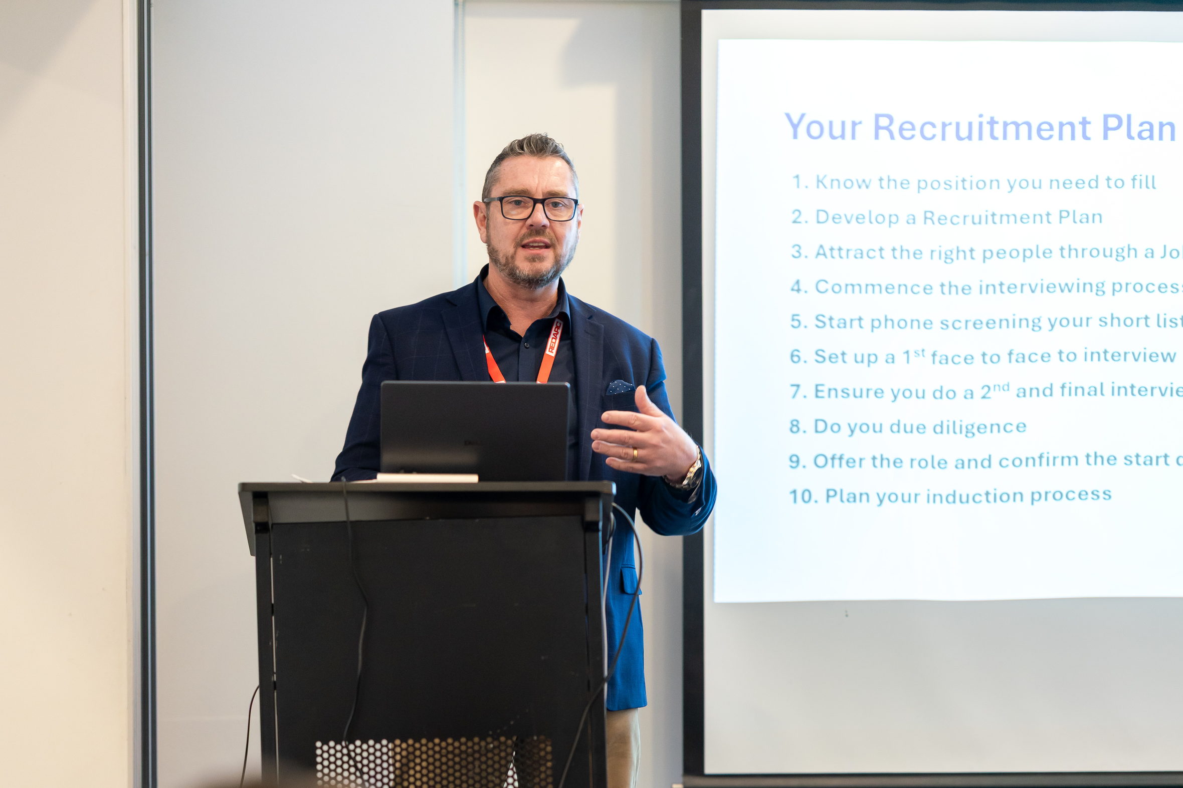 Colin Wilson at a lectern walking a business audience through a ten-step recruitment plan on screen